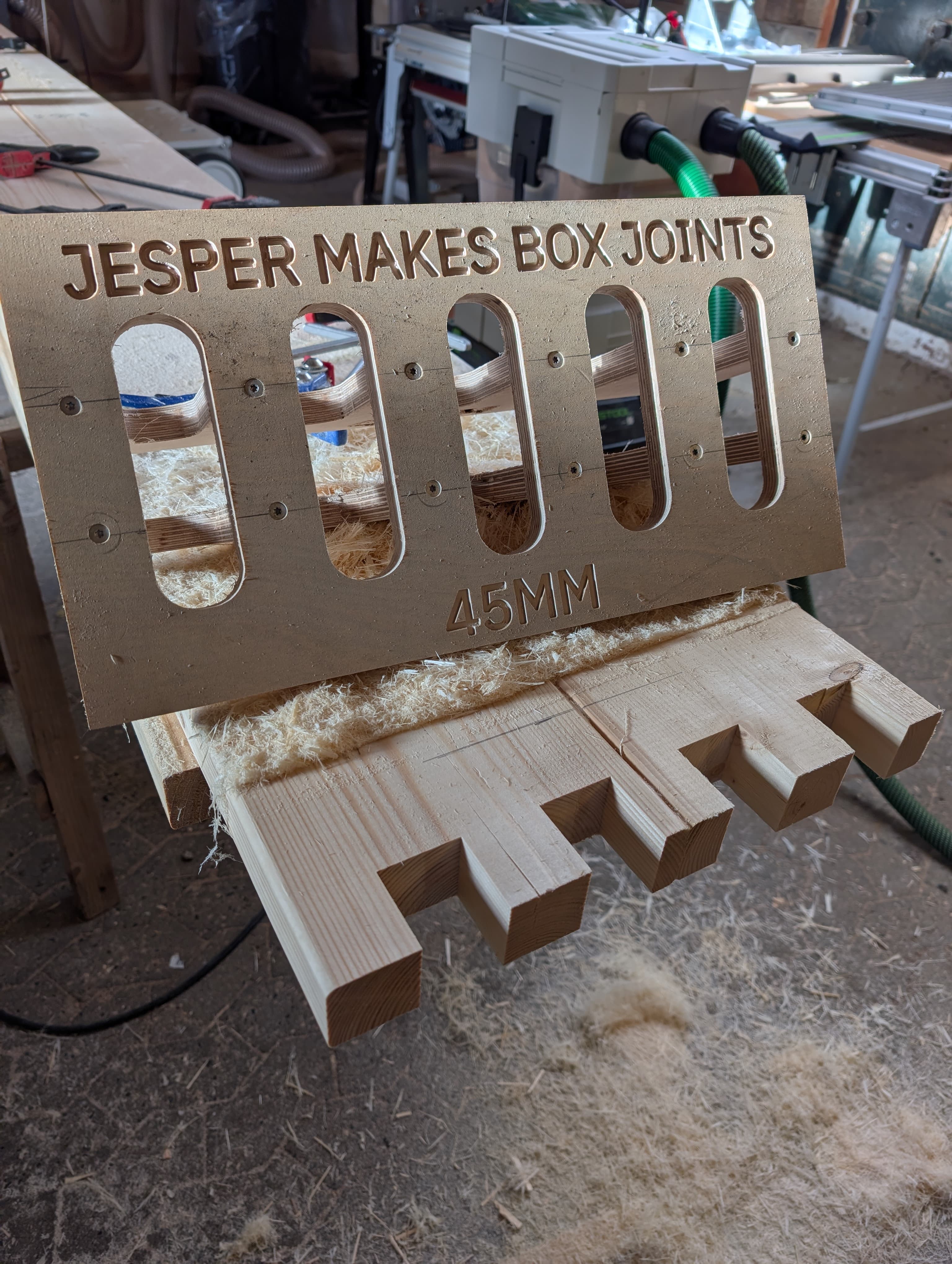 A 21mm birch plywood routing template labelled 'JESPER MAKES BOX JOINTS 45MM' sitting on top of a fir board with freshly routed finger joints